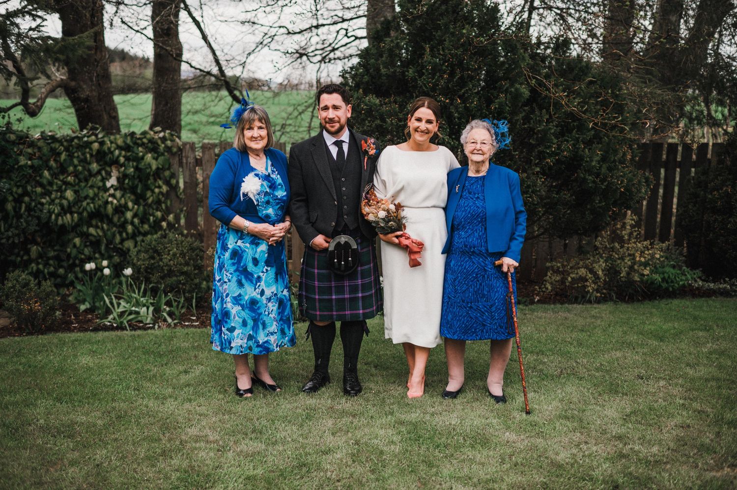 Family group photo at Scottish wedding with traditional kilts and formal attire in garden setting.