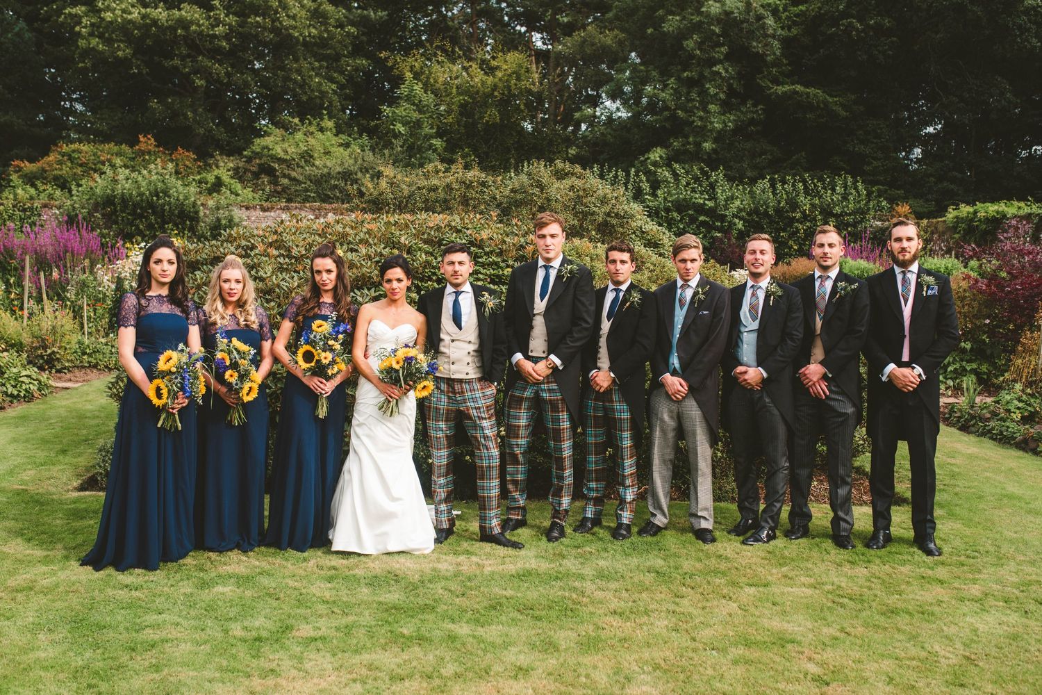 Formal wedding party line-up with navy bridesmaids dresses and Scottish kilts in garden setting.