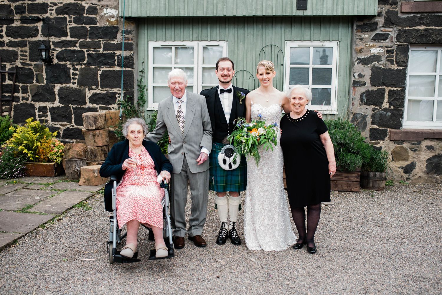 A small wedding party poses in front of a stone building with a green door, featuring traditional Scottish attire.
