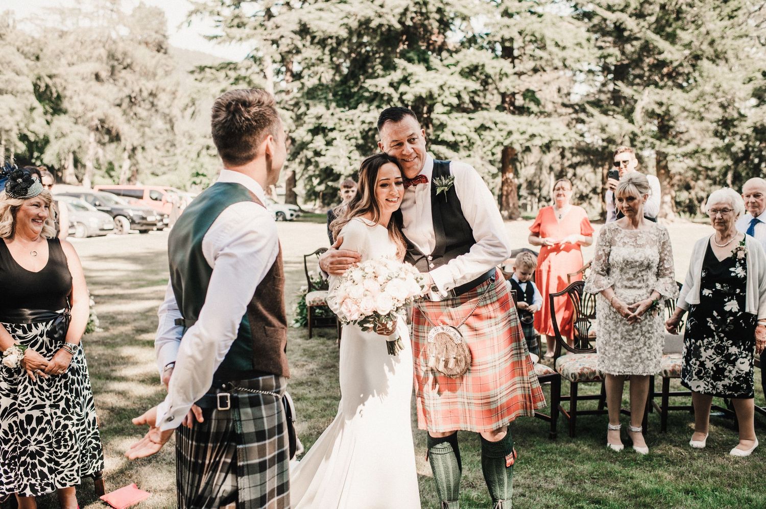 Wedding ceremony outdoors with guests in traditional Scottish kilts and formal attire.