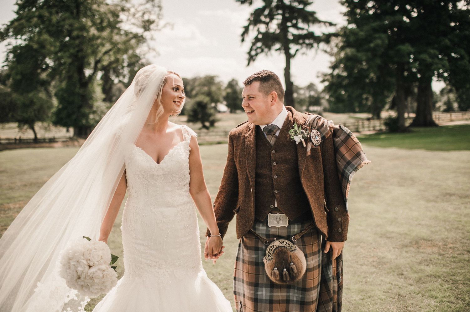 Romantic wedding couple poses outdoors with bride in lace gown and groom wearing traditional Highland dress.