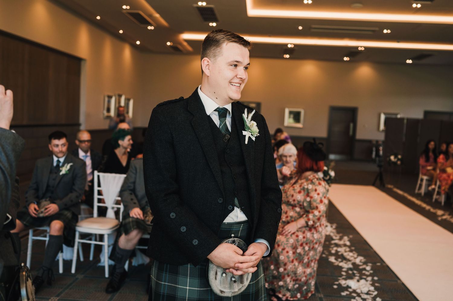 Groom in traditional Scottish kilt and formal jacket stands during wedding ceremony.