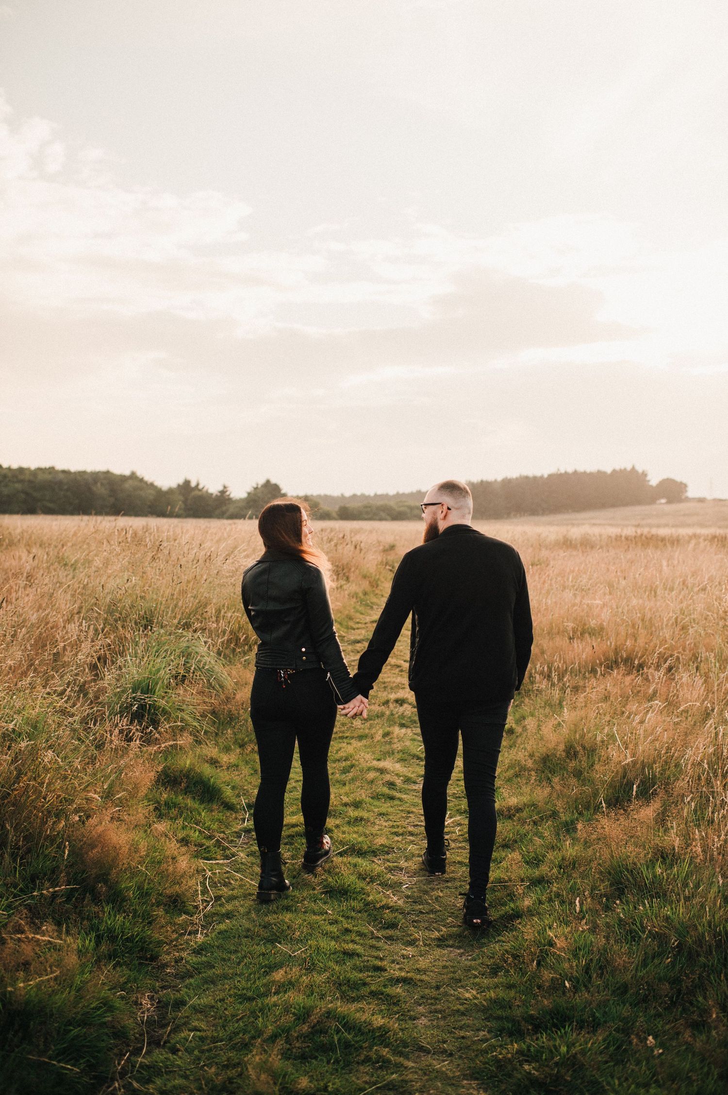 Two figures holding hands while walking through a sunlit meadow with forest in the distance.