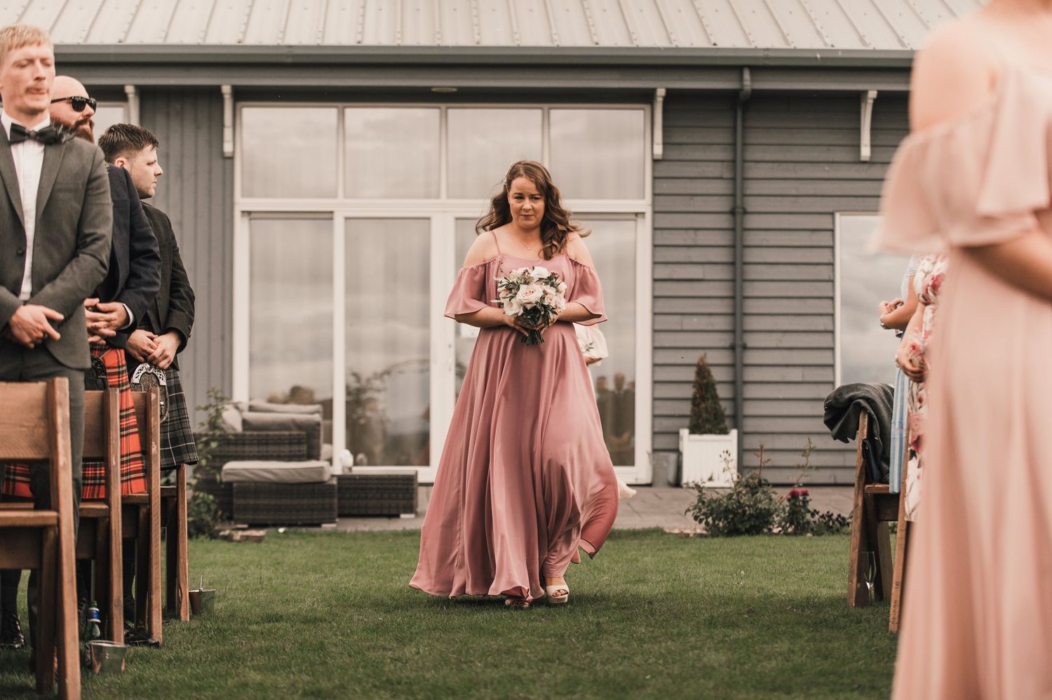 Bridesmaid in a flowing pink gown walks down outdoor ceremony aisle with modern building backdrop.