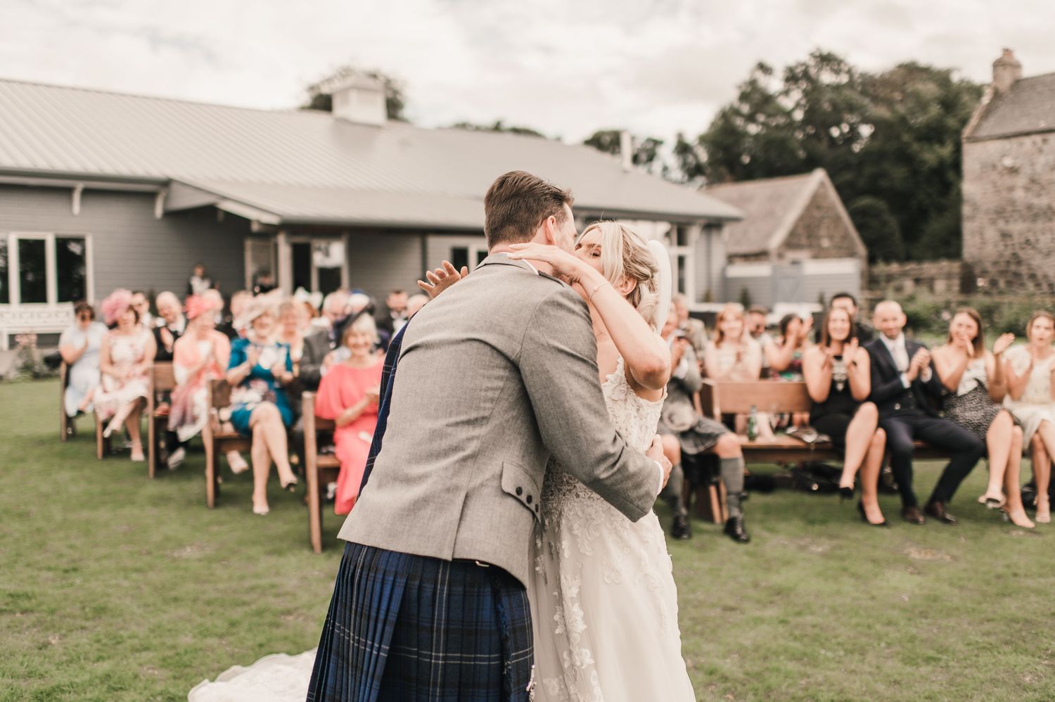 A romantic first kiss moment during an outdoor wedding with seated guests in the background.