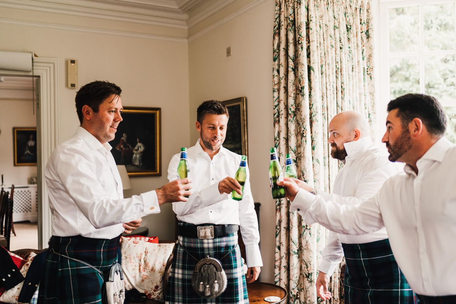 Groomsmen in white shirts and kilts toast with drinks in an elegant room.