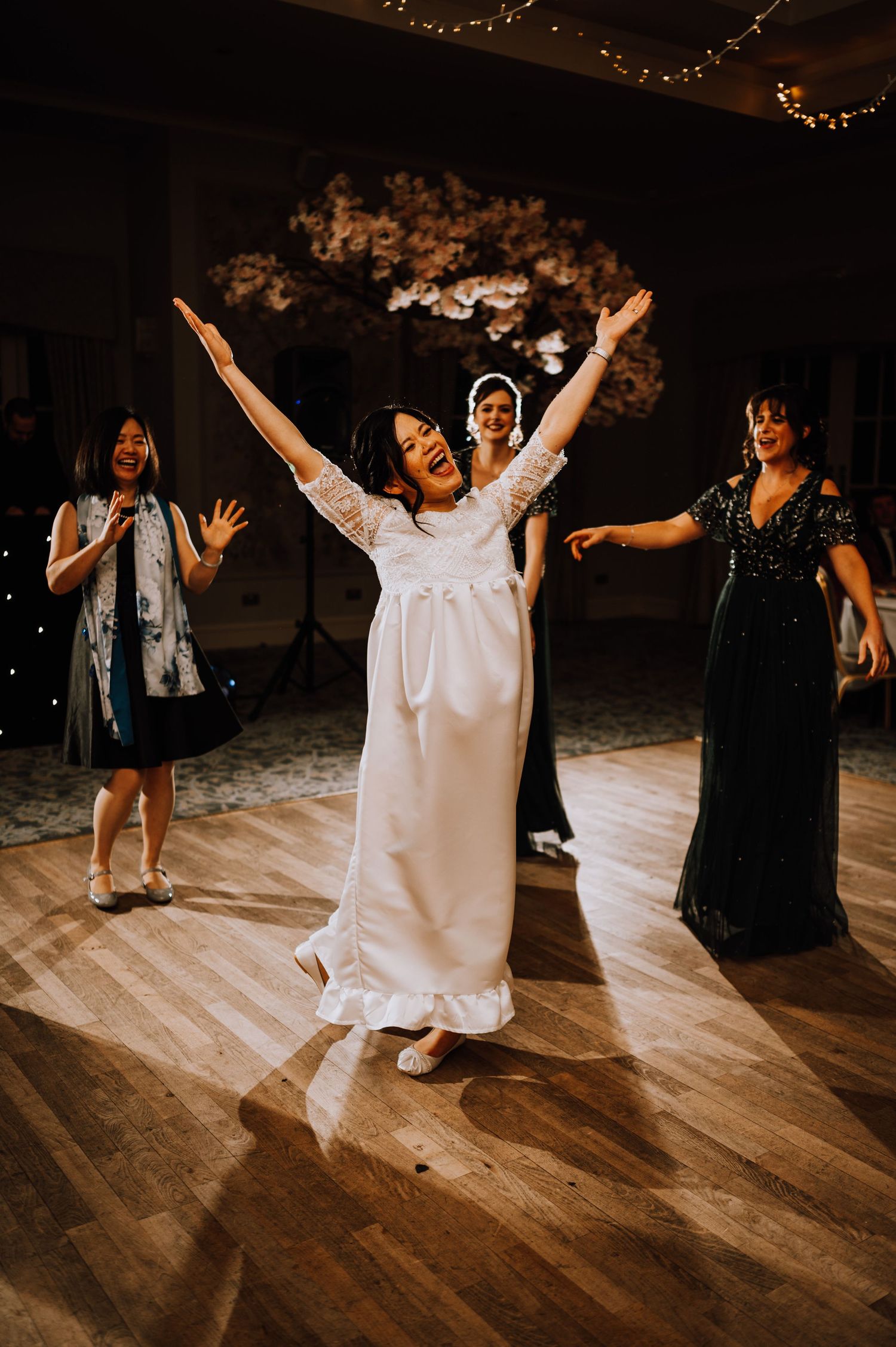 A person in a white wedding dress dances enthusiastically with raised arms on a wooden dance floor.