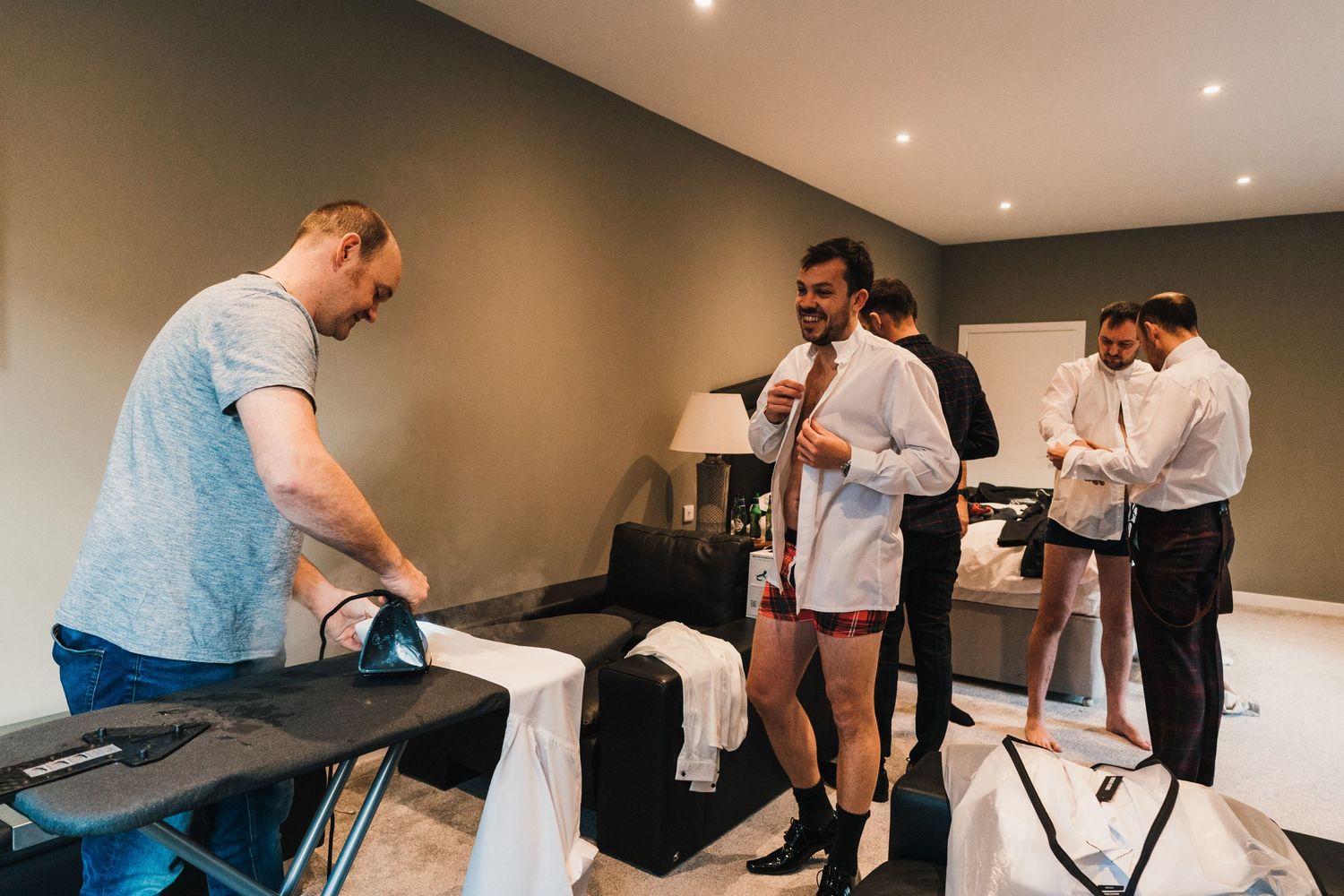 Groomsmen getting ready in a hotel room while checking their phones and relaxing before the wedding ceremony.