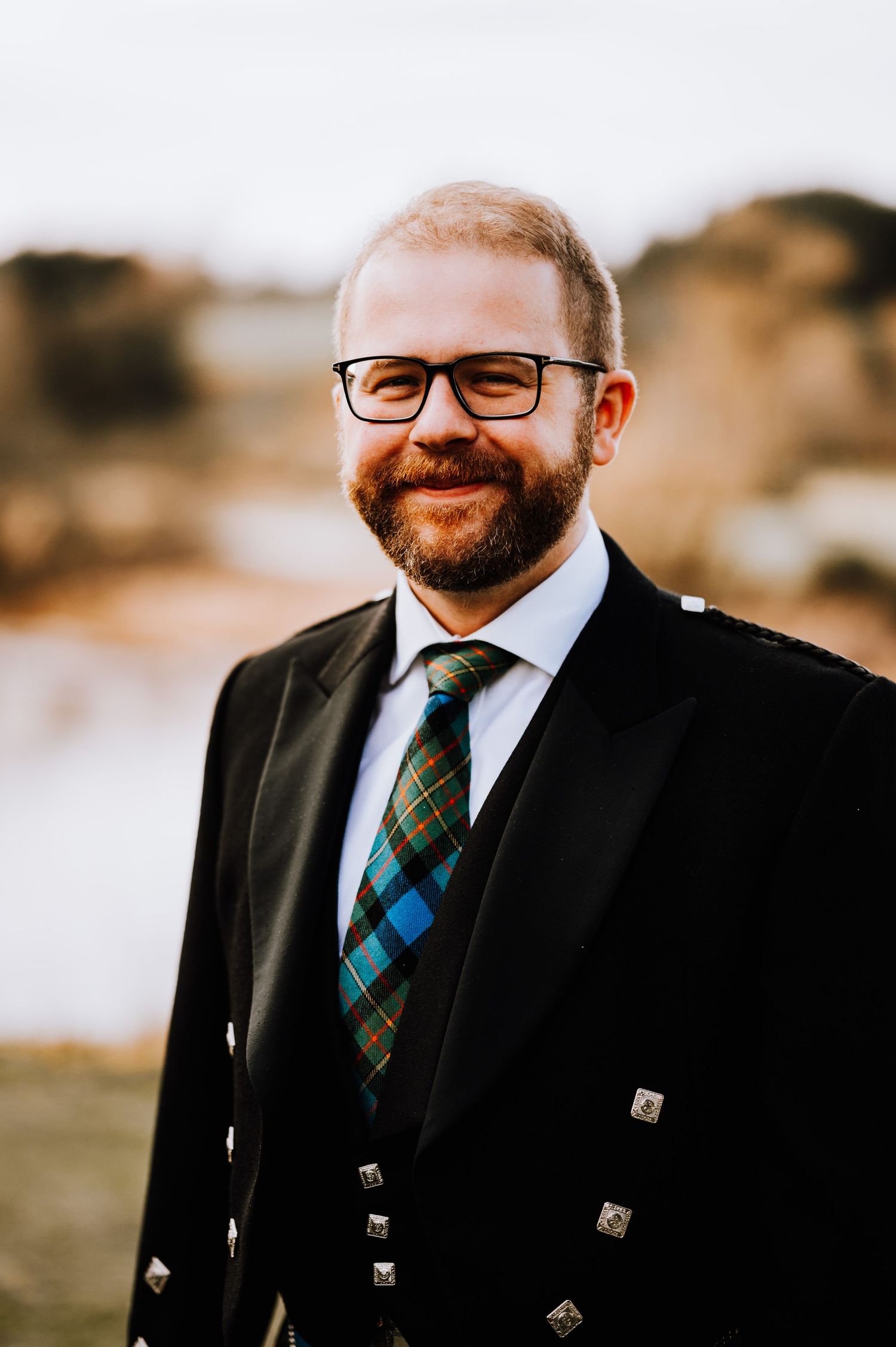 Person in formal Scottish attire with plaid tie and black jacket poses outdoors.