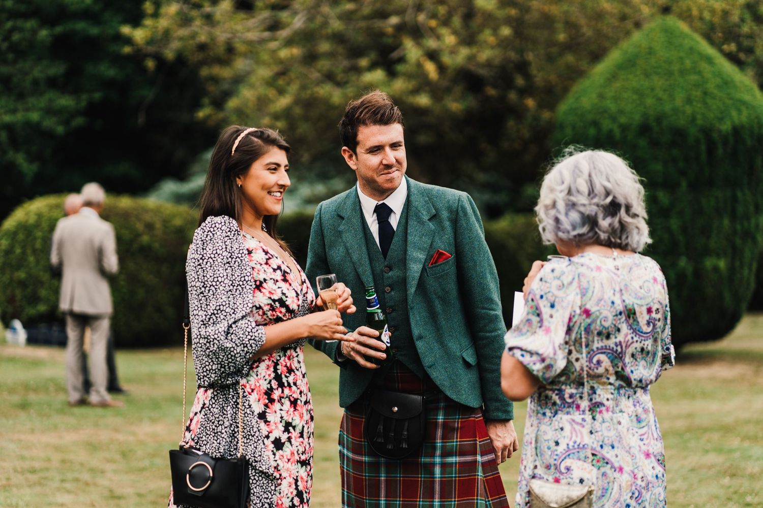 Guests in suits and formal wear mingle at outdoor wedding reception.
