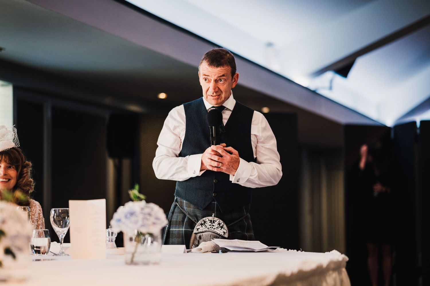 A speaker in formal attire addresses guests at a wedding reception.