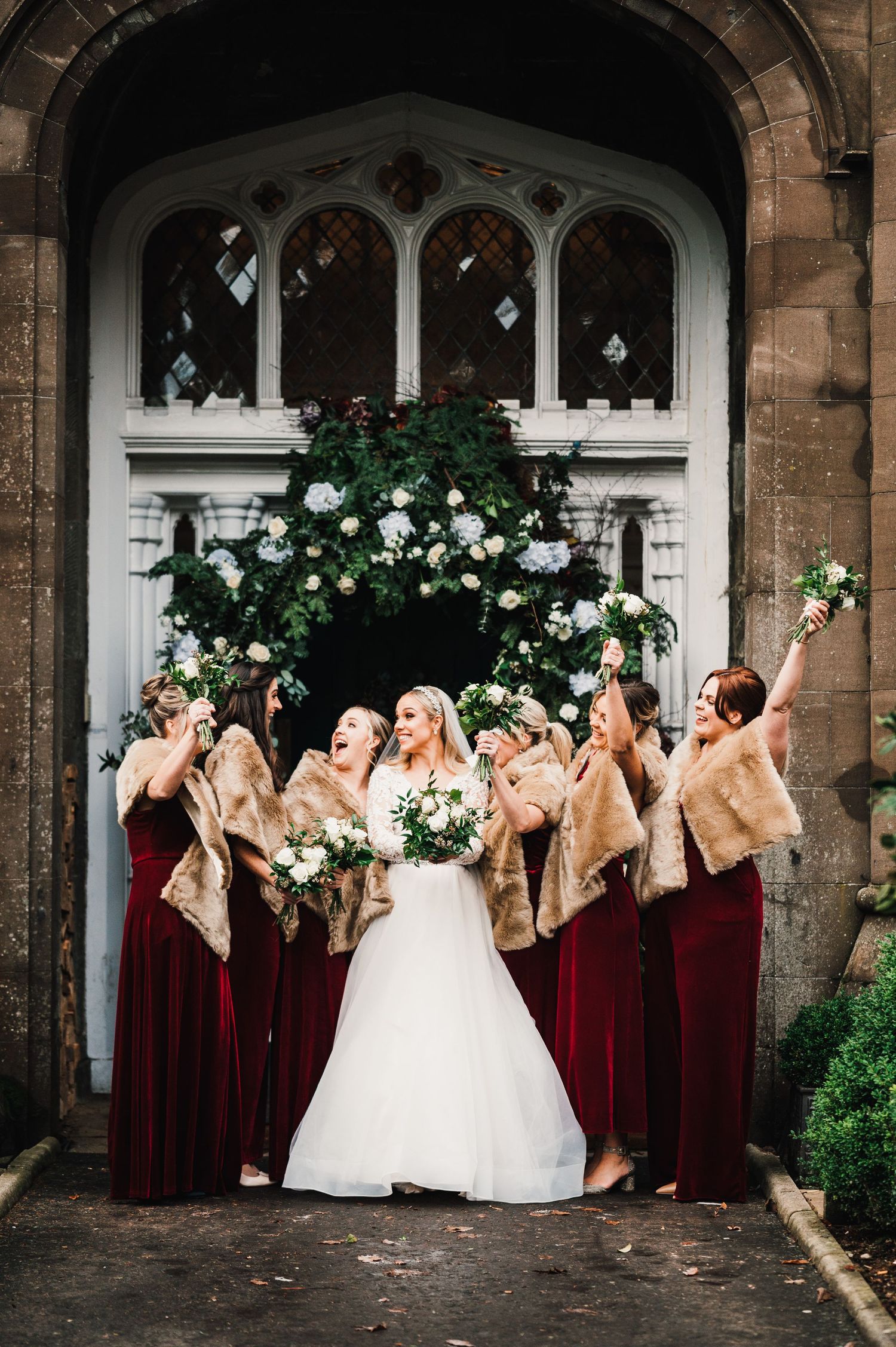 Bridesmaids in burgundy dresses share a joyful moment with the bride under a Gothic archway.