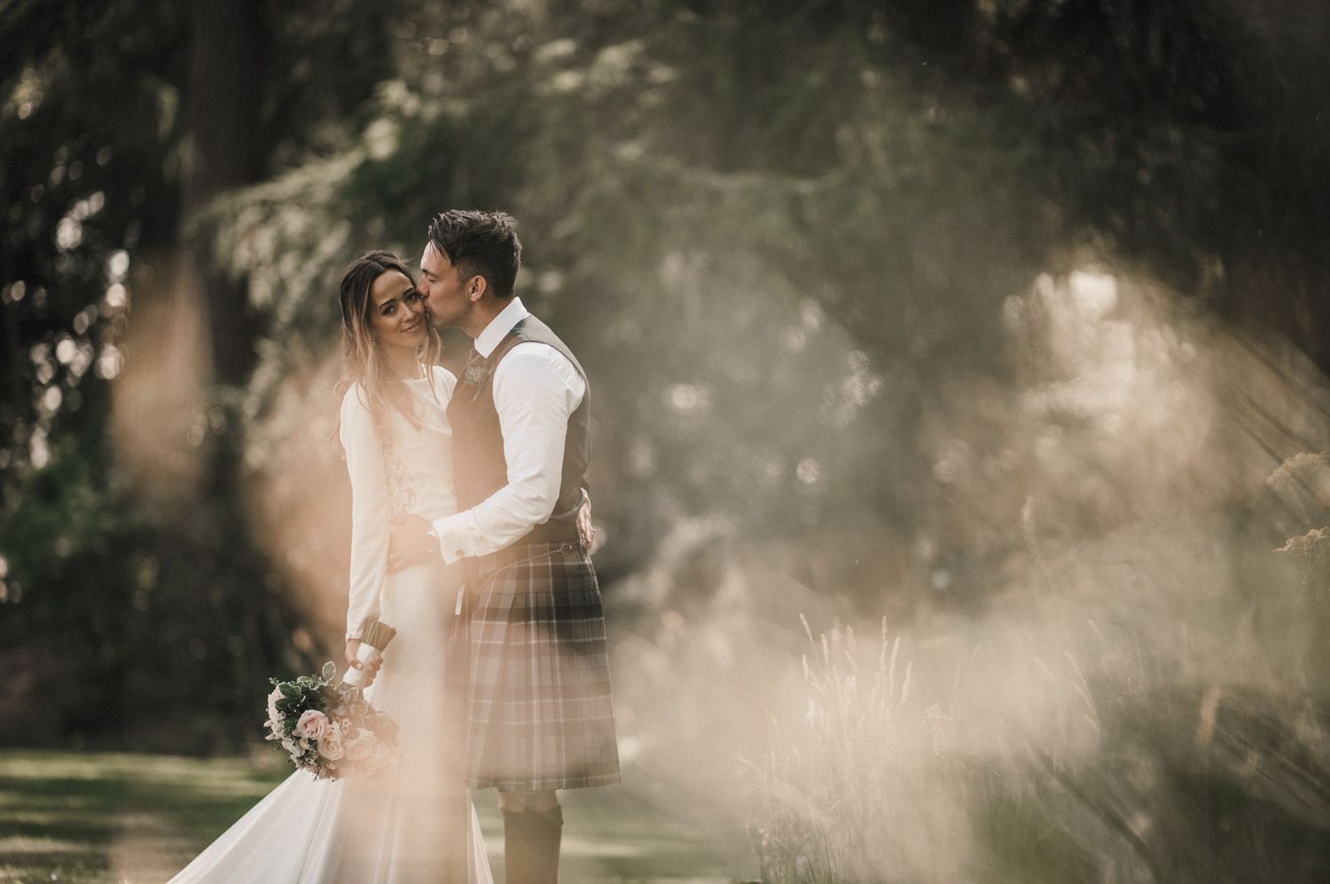 Ethereal wedding moment with sunlit mist surrounding couple in Scottish-themed outdoor ceremony.