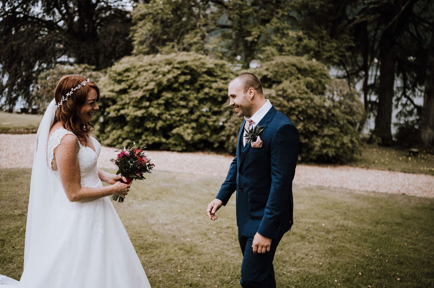Couple in wedding attire stand on grassy area with trees and water in background.