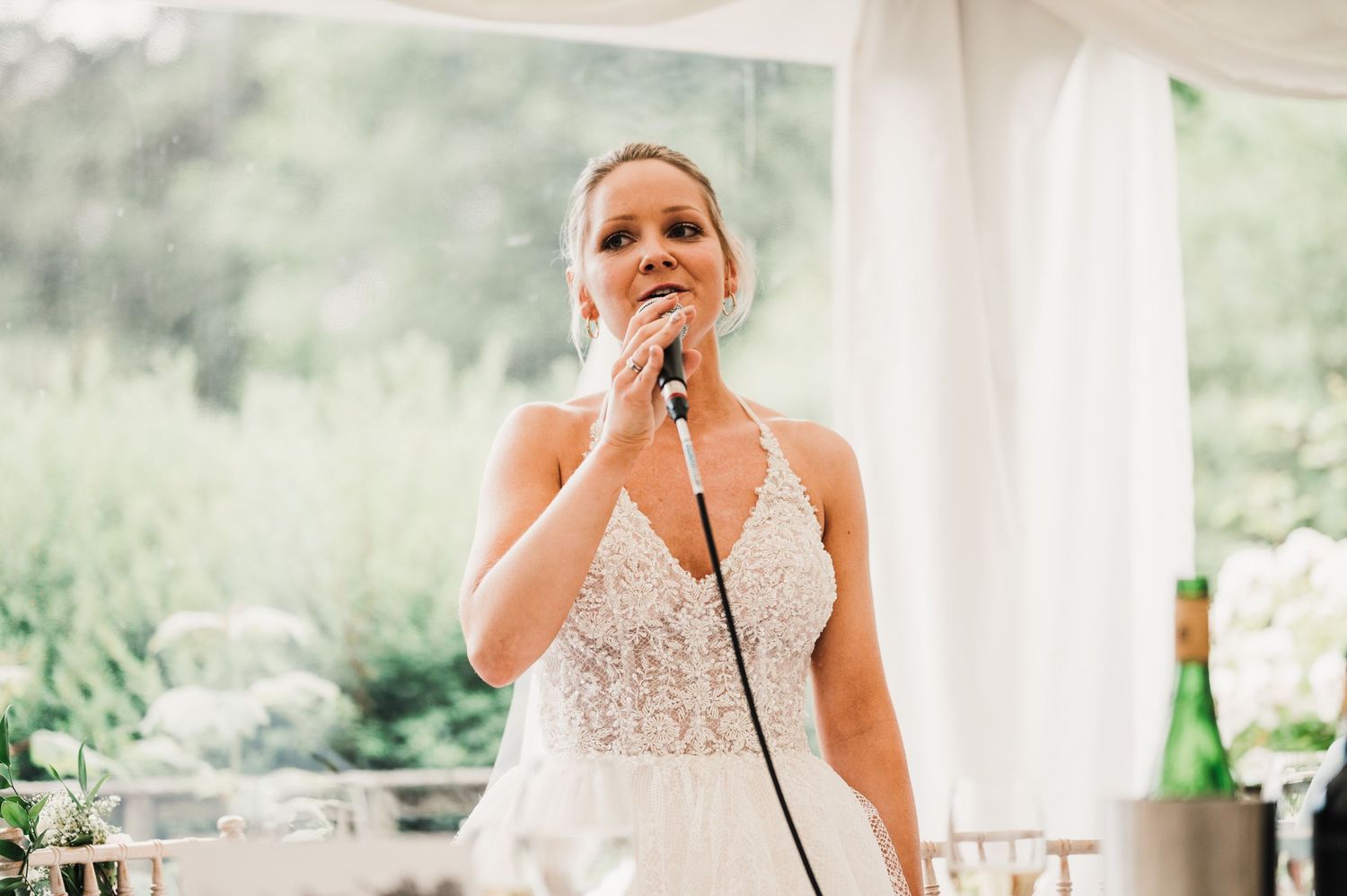 Wedding speech being delivered under white tent with elegant decor and natural scenery visible outside.