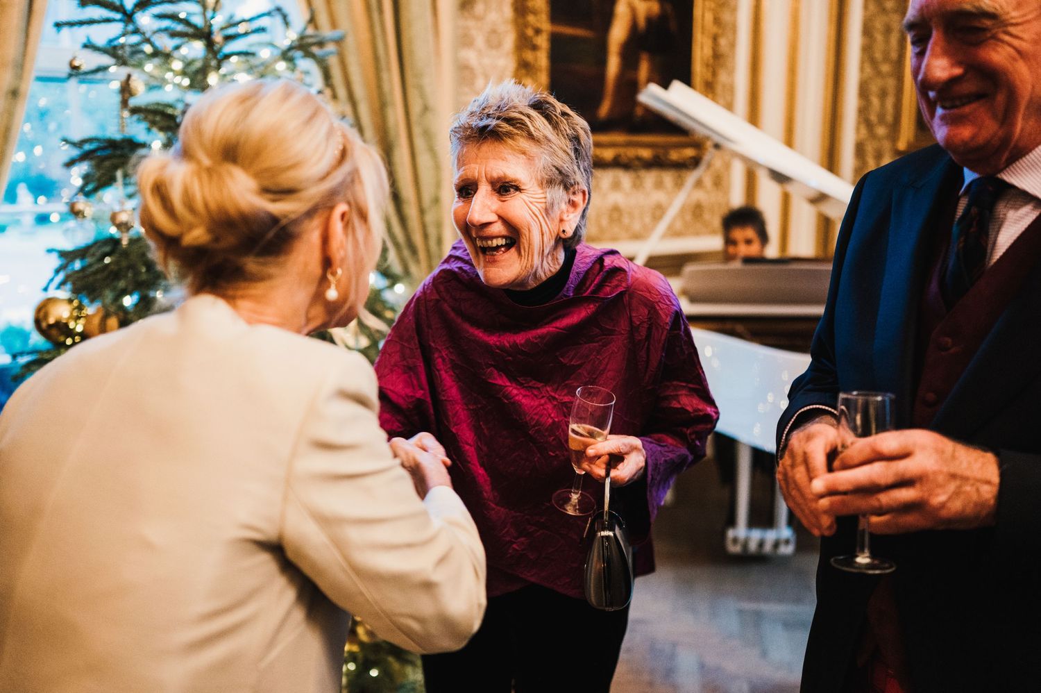 Guests share a lively conversation beside a decorated Christmas tree at an elegant indoor party.