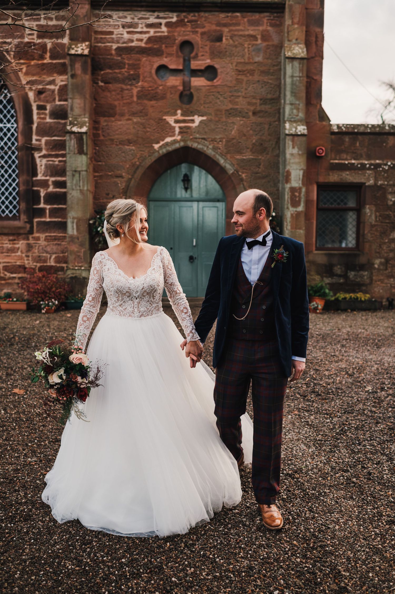 Newlyweds stand hand-in-hand outside historic brick church with gothic arched doorway.