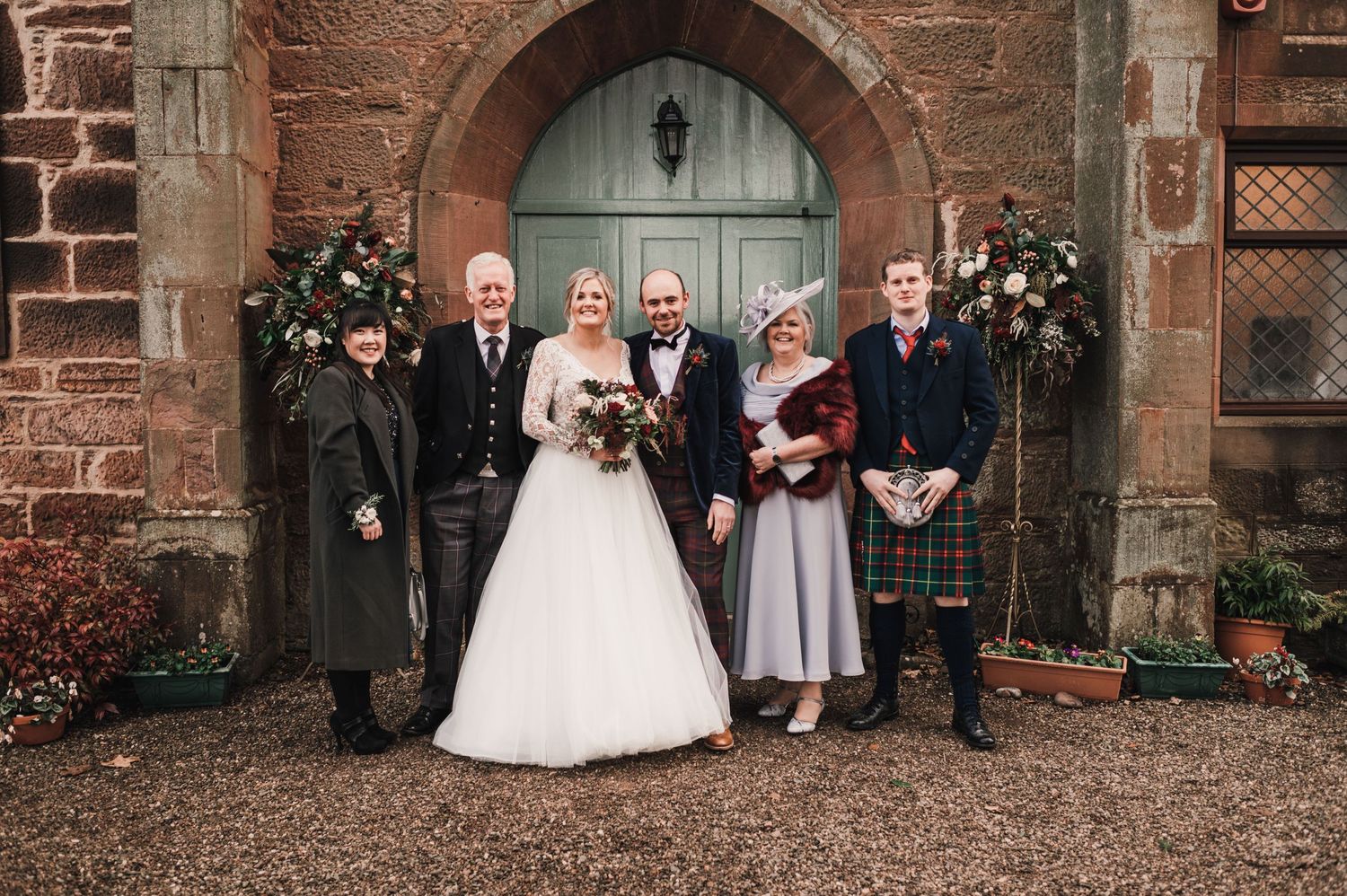 Small wedding group poses in formal attire by church entrance with floral decorations.
