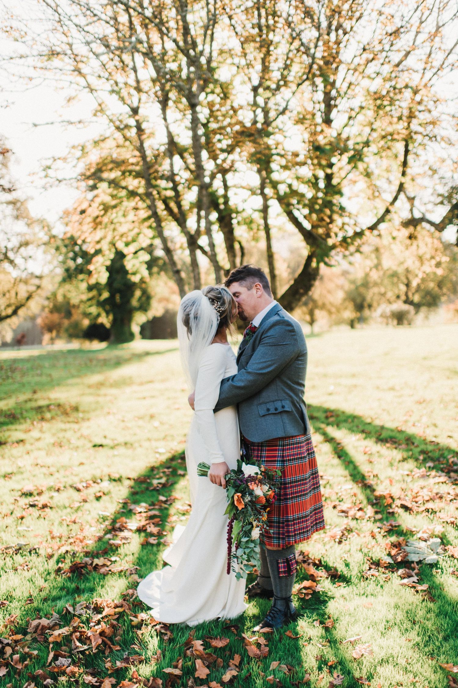 Wedding couple in Scottish attire embracing in an autumn garden with golden sunlight filtering through trees.