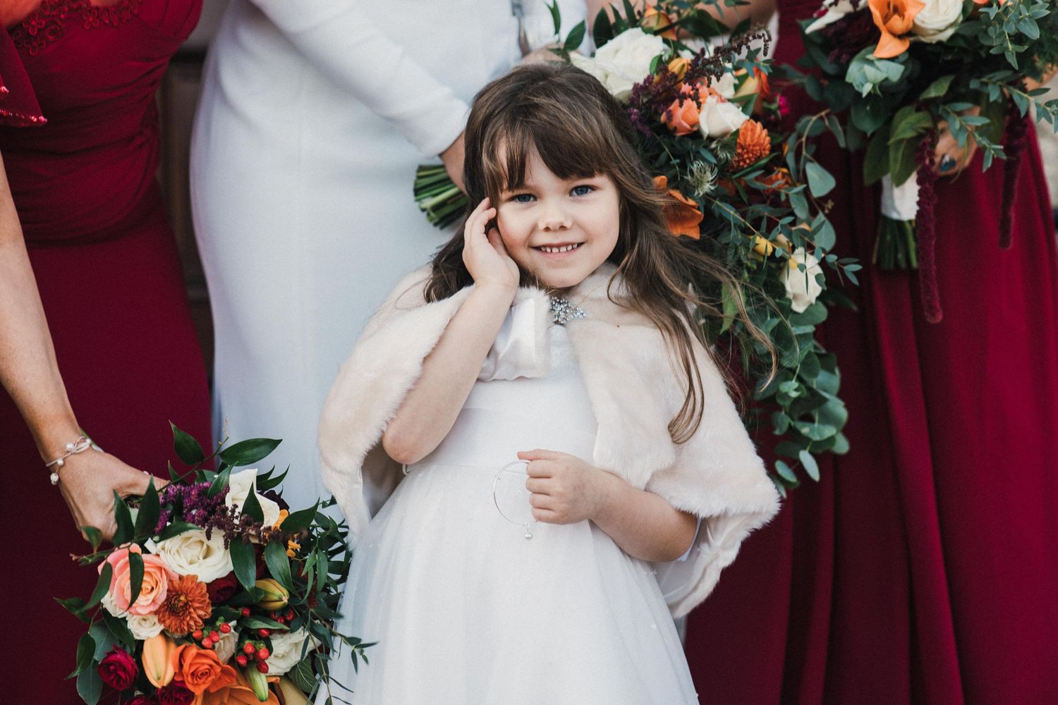 A young flower girl in white dress poses with floral arrangements and burgundy decor.