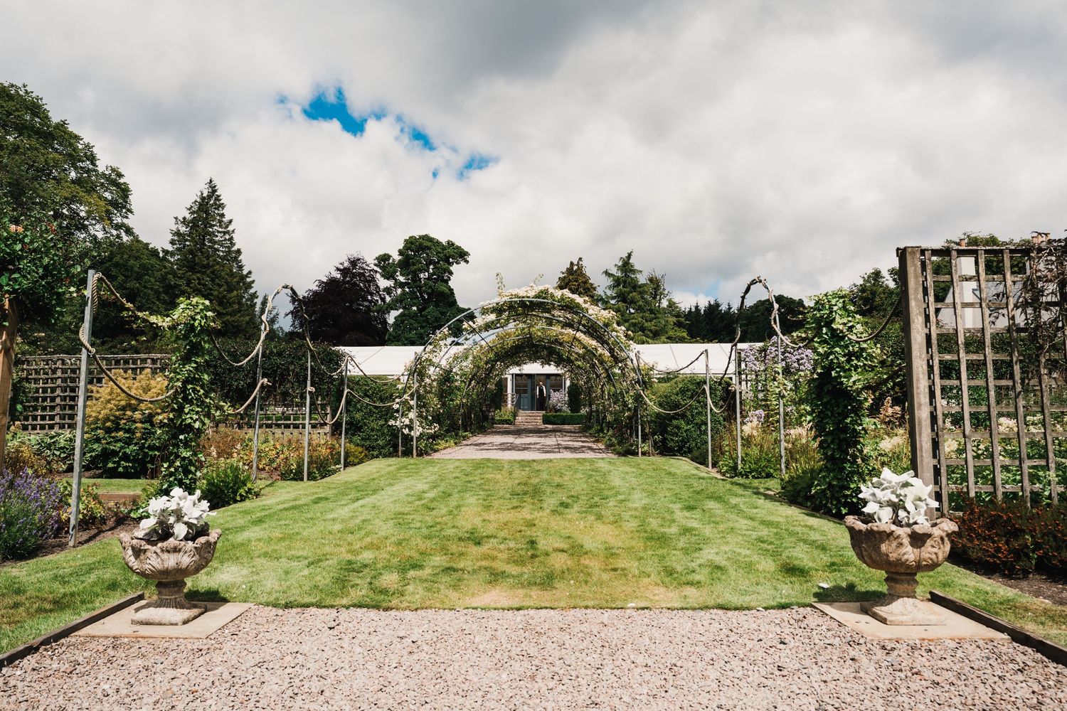 Manicured garden wedding venue with stone pathway and floral arch under cloudy skies.
