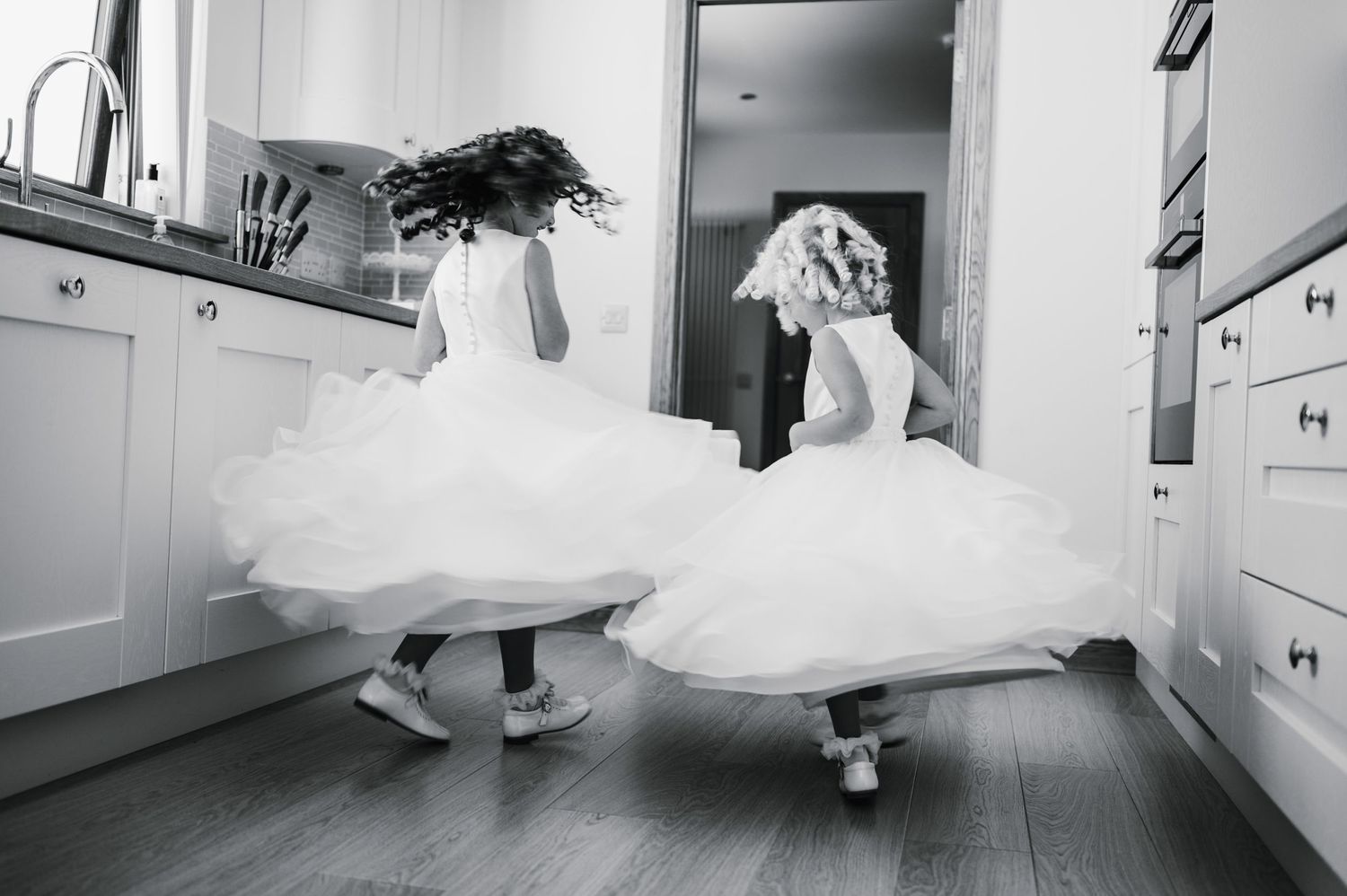 Black and white photo of two attendants twirling in white dresses in a kitchen.