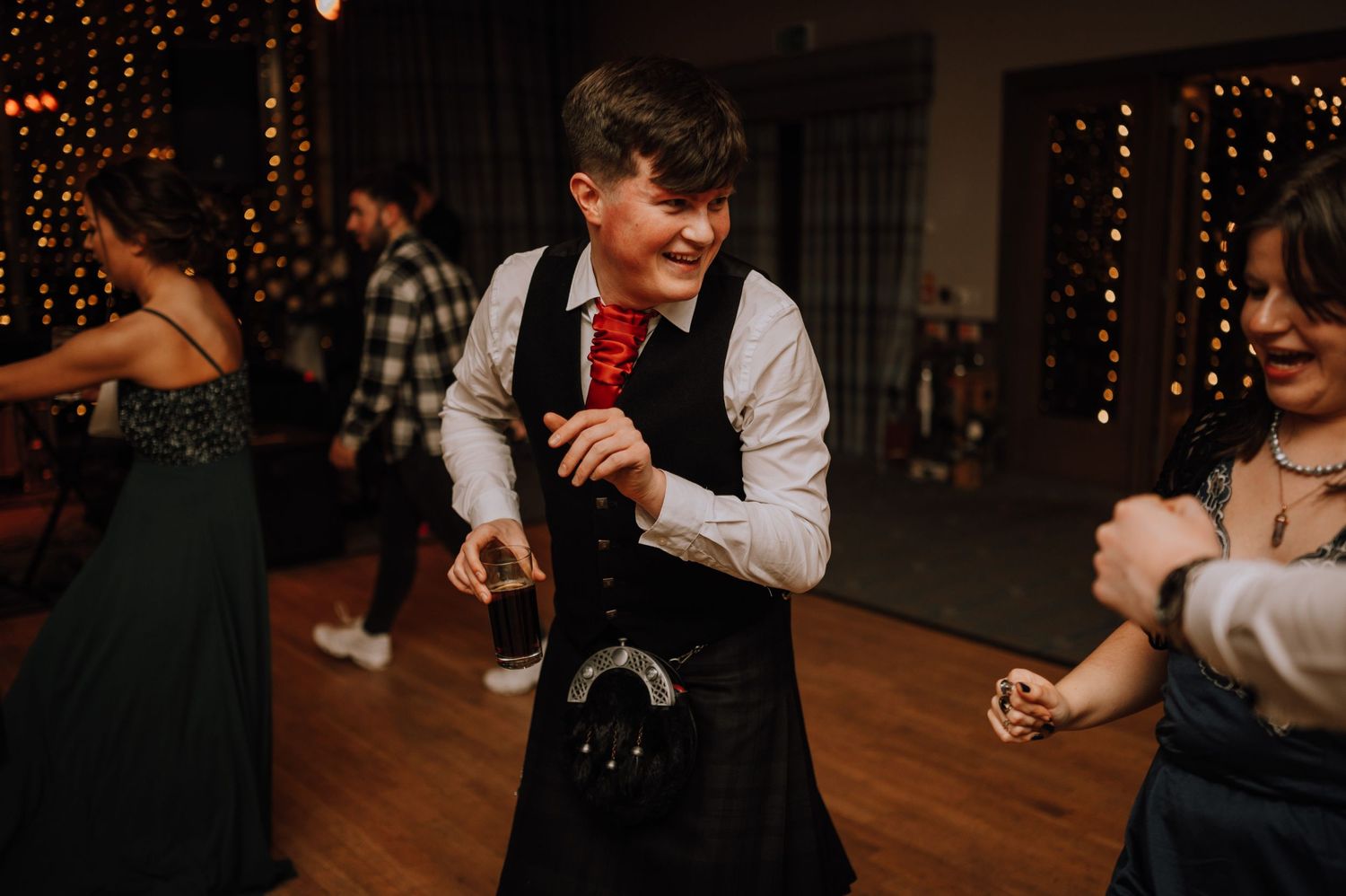 Guests dance and celebrate on a wooden floor under string lights at an indoor venue.