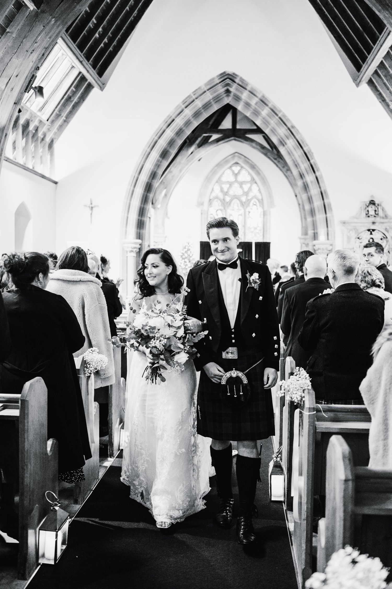 Couple walks down church aisle after ceremony, groom in traditional Scottish kilt.