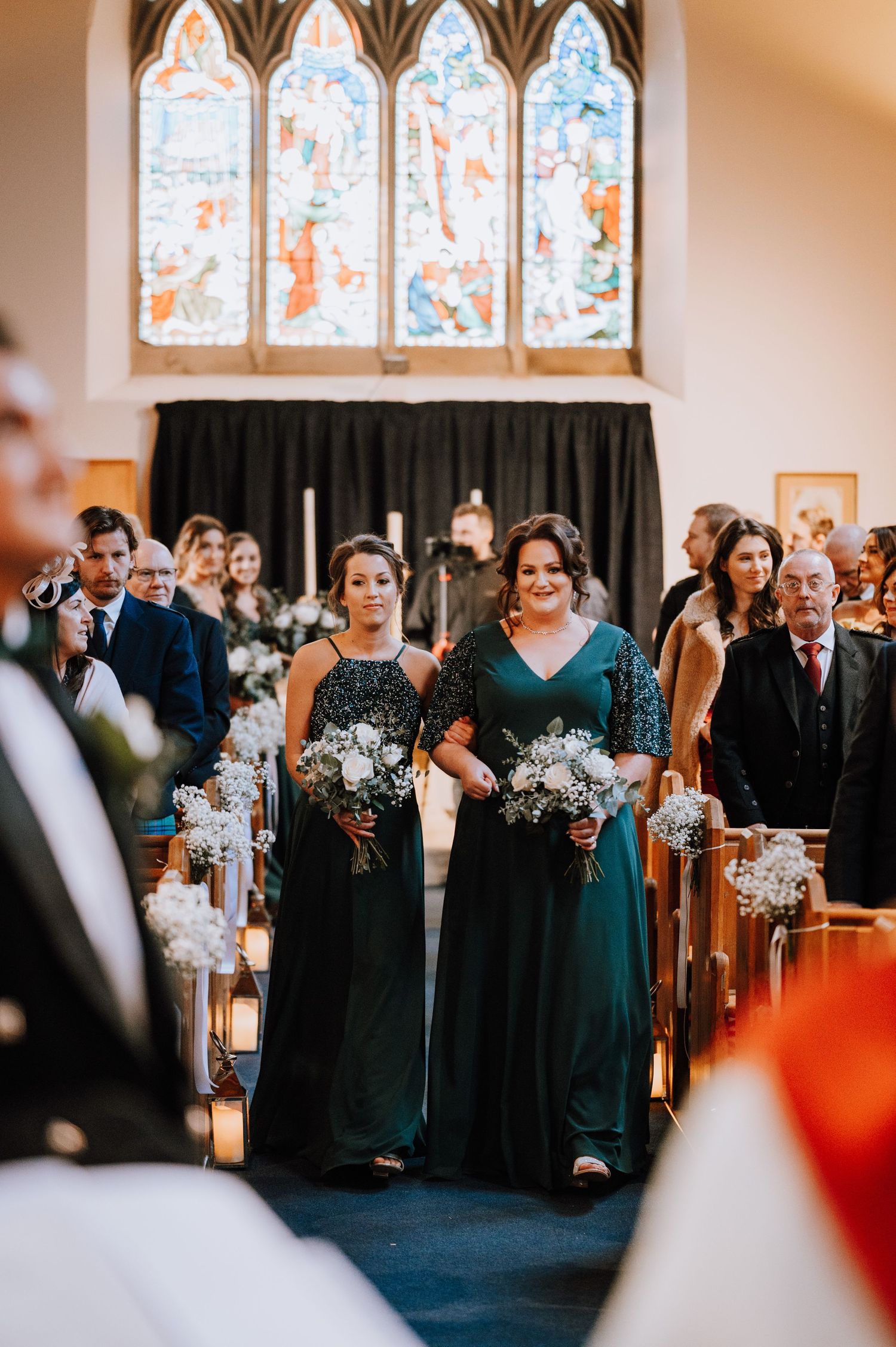 Bridesmaids in dark green dresses walking down church aisle with stained glass windows visible.