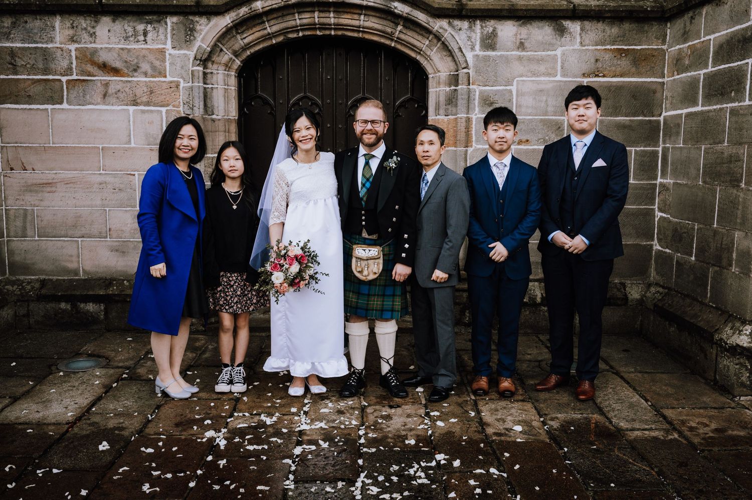 A wedding party poses in front of a stone archway with the groom wearing traditional Scottish kilt attire.