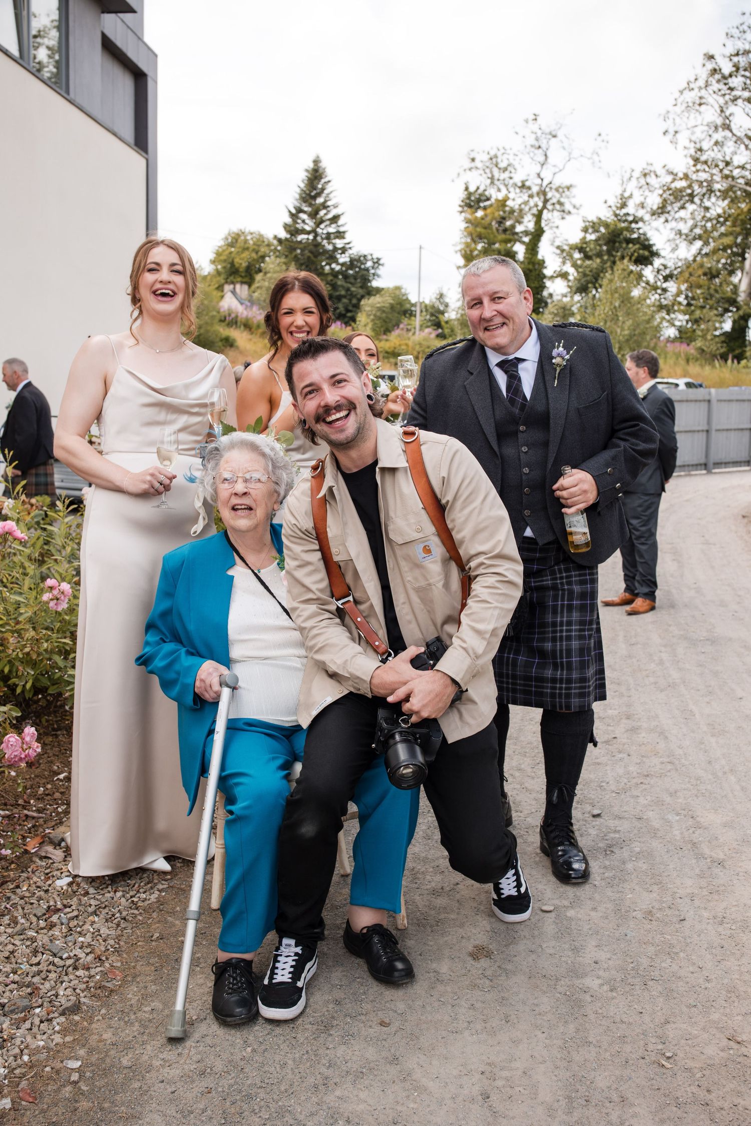 A group of wedding guests pose together on a path outdoors with trees and flowers in the background.