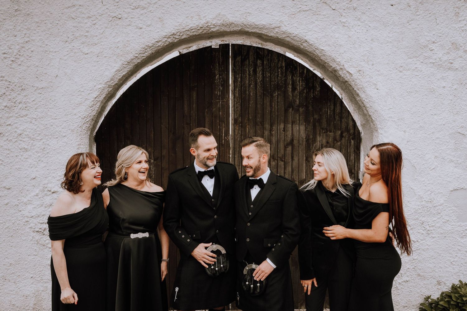 Logie House Wedding Photography. Wedding party in formal black attire posing against white stone archway.