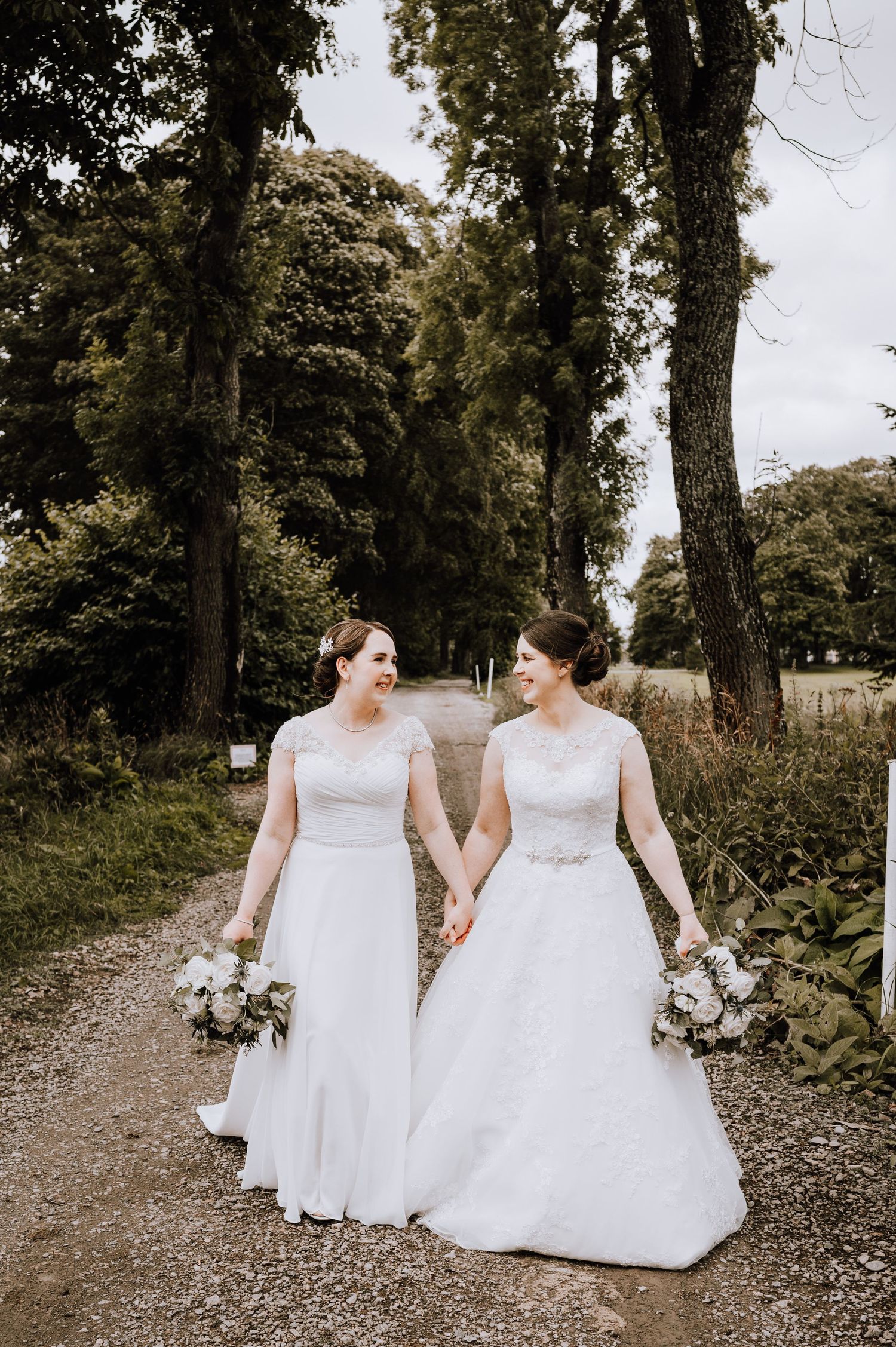 Two brides in white wedding dresses walking hand in hand on outdoor path Meldrum House Wedding Photography.