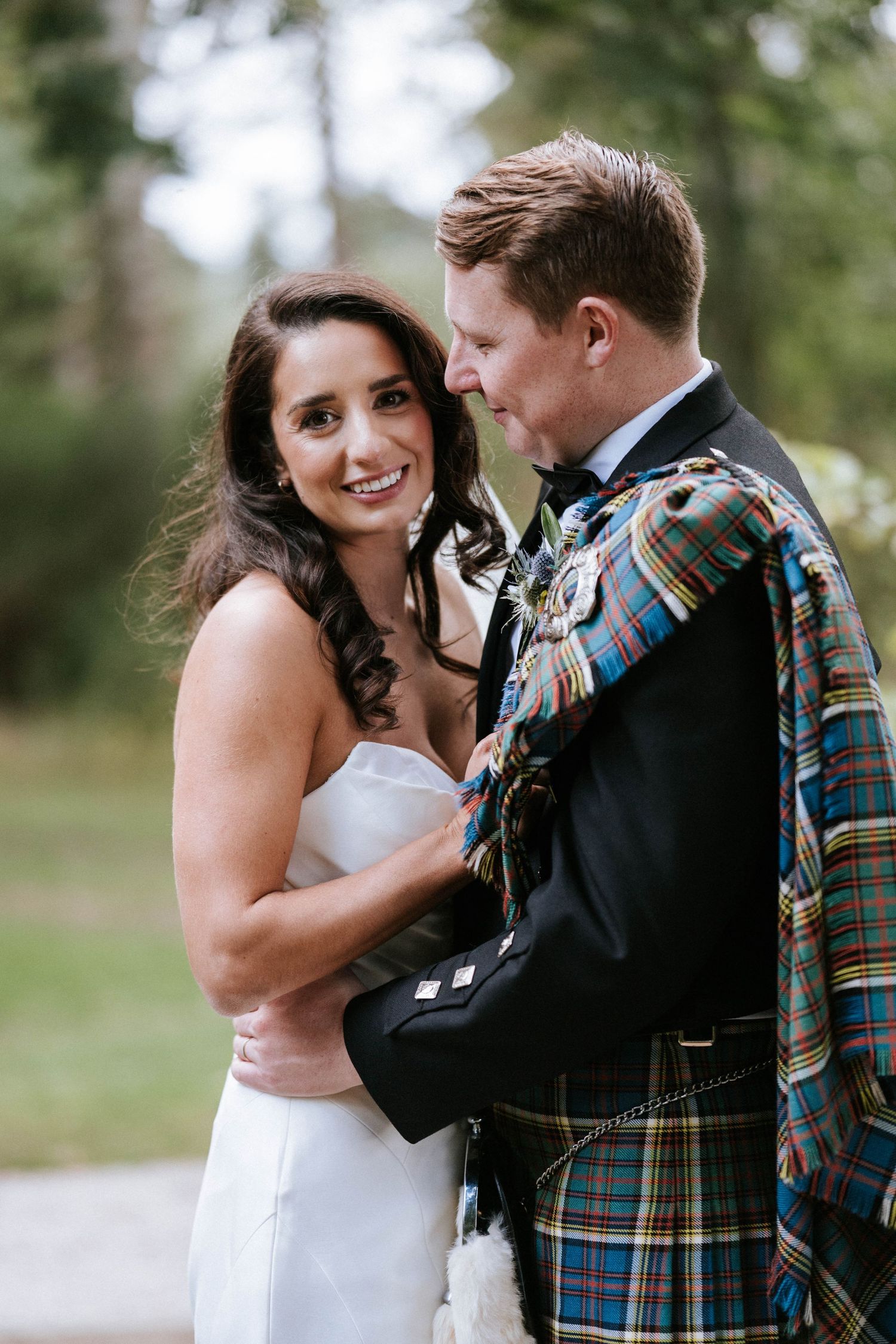A romantic wedding portrait featuring traditional Scottish attire against a blurred natural backdrop.