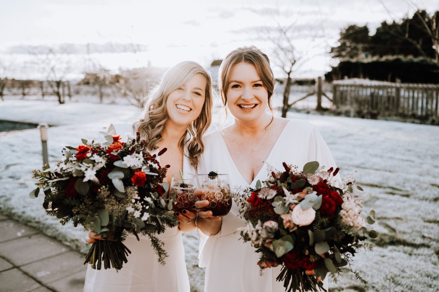 Same Sex Wedding Photography. Two people hold burgundy and white floral bouquets while smiling in a snowy winter setting.