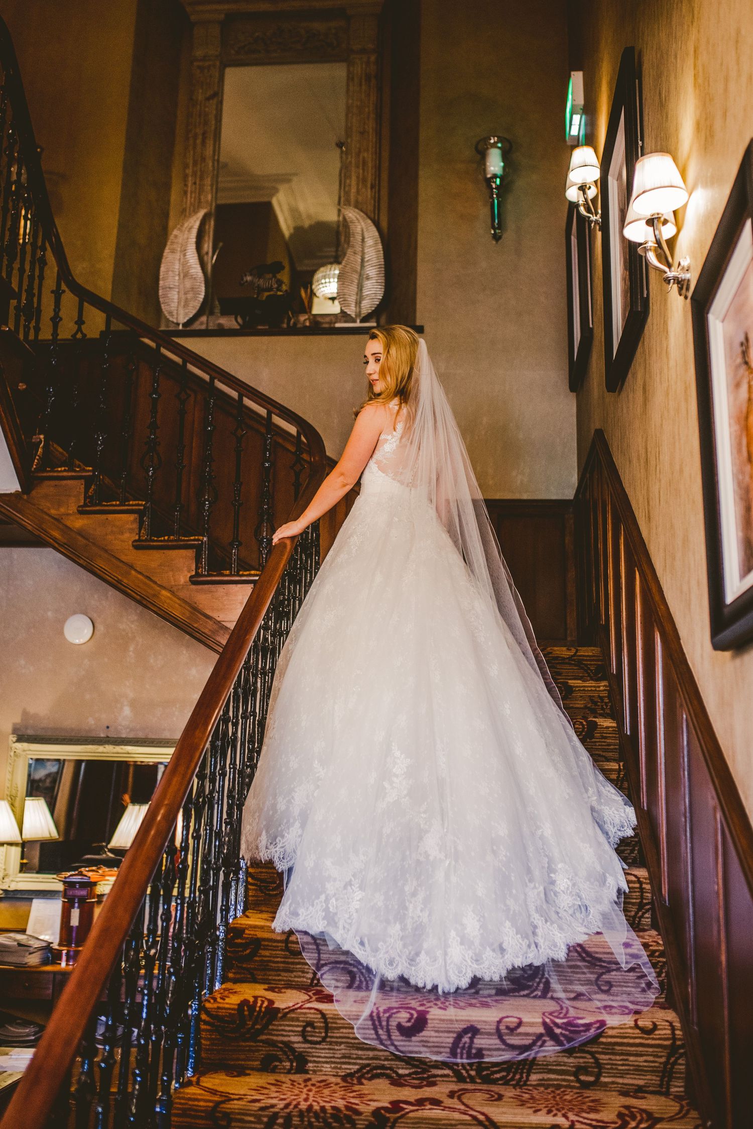 A bride in a flowing white lace wedding gown with long veil stands on an elegant staircase with ornate iron railings.