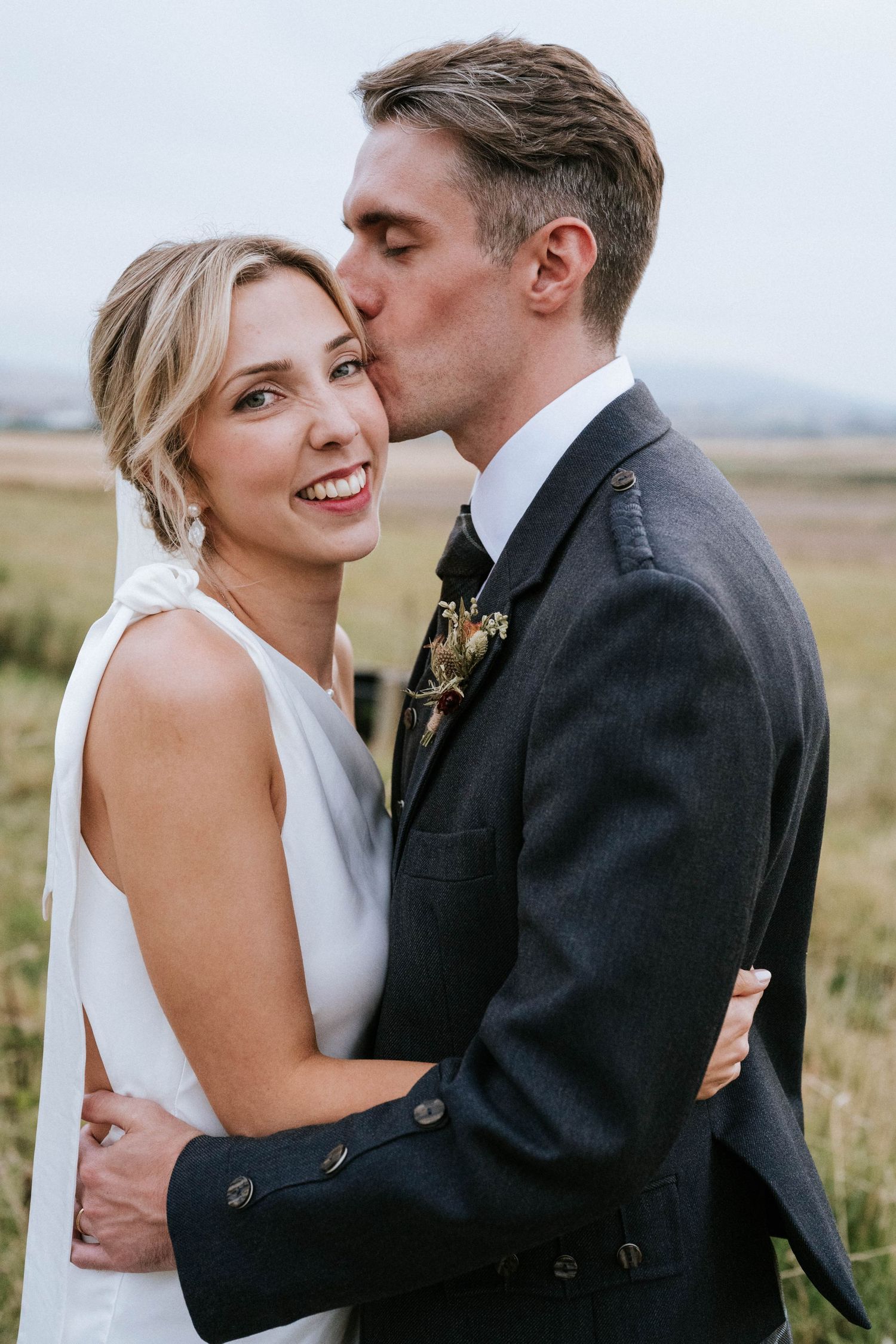 A couple embraces in an intimate moment against a backdrop of open fields.