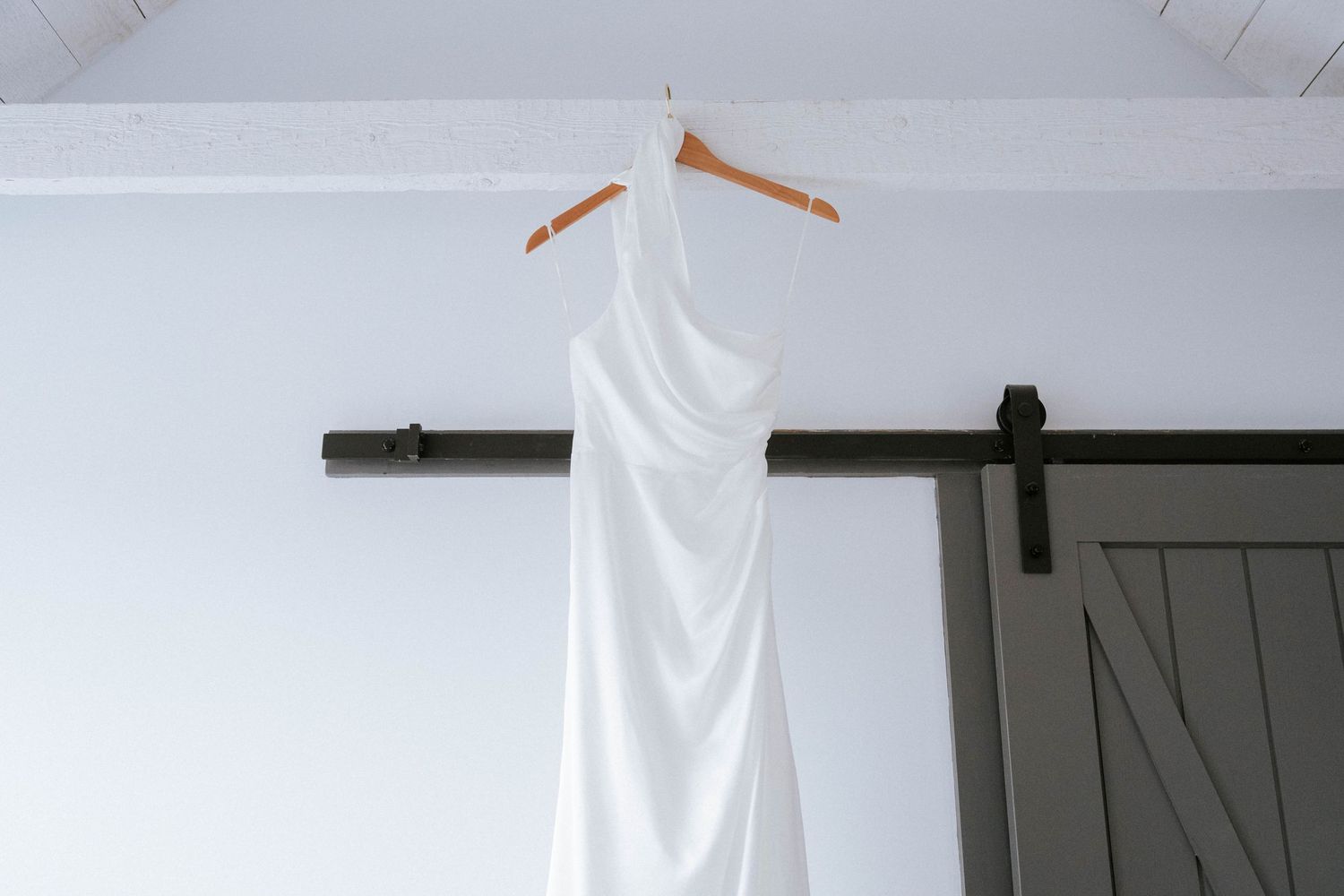 Elegant white wedding dress hanging on wooden hanger against grey barn door.