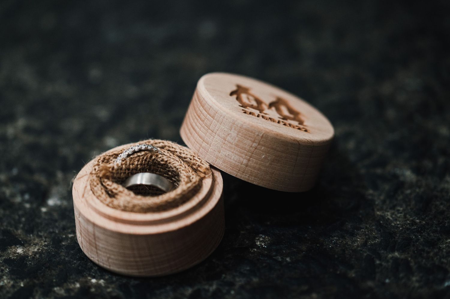 Two wooden ring boxes with carved designs sit on a dark textured surface in moody lighting.