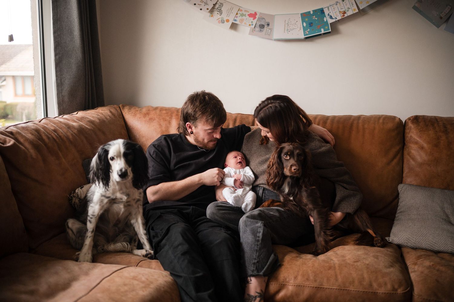 Family relaxing on brown leather couch with dog and baby in casual home setting.