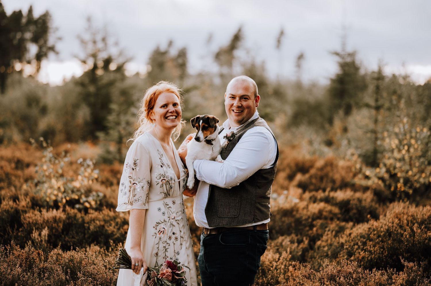 A happy couple poses with their small dog in a scenic heather field.