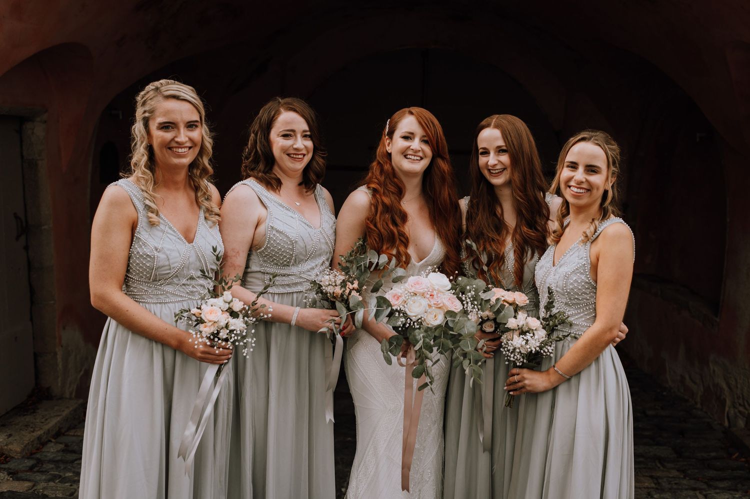 Bridesmaids in elegant silver grey dresses holding floral bouquets pose together under stone archway.