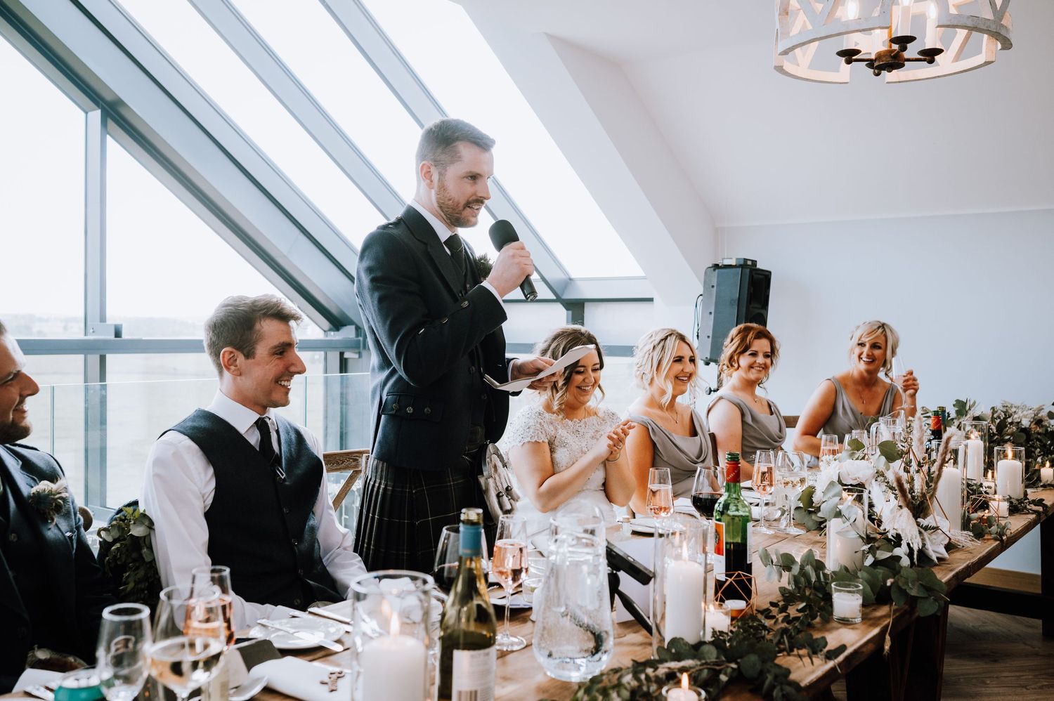 Best man delivers a speech at wedding reception while guests listen attentively.