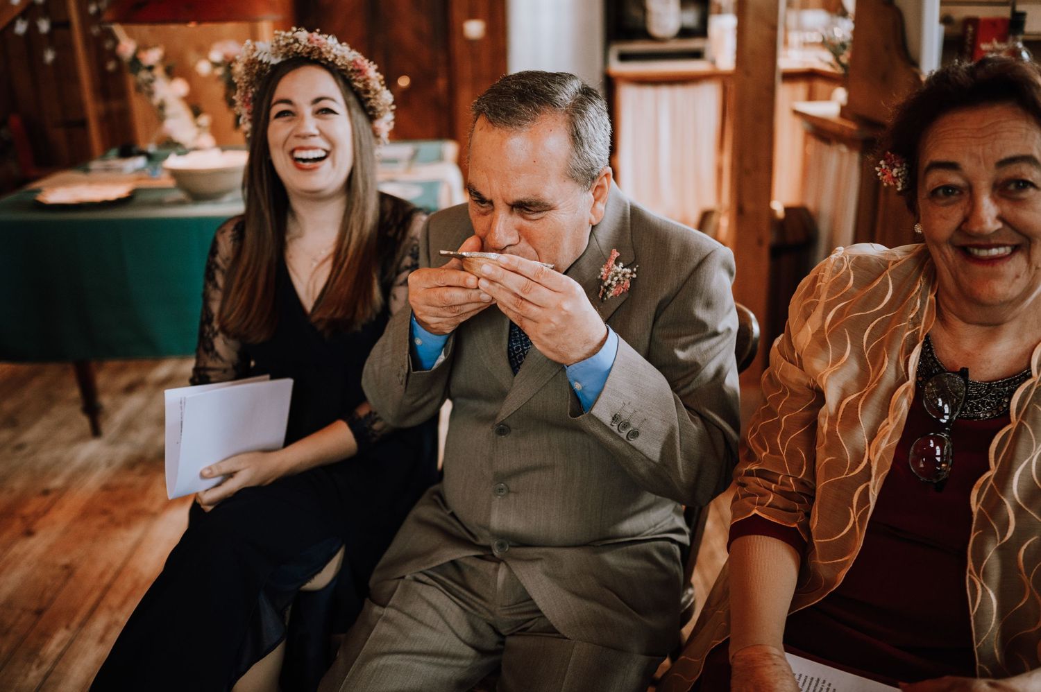 Wedding guests laugh and react emotionally during a ceremony in a rustic wooden interior setting.