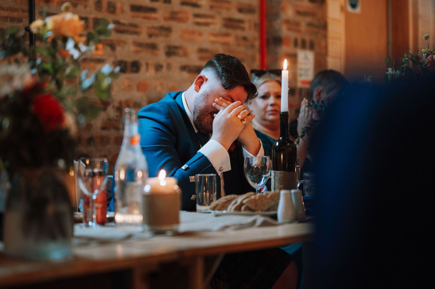 A guest rests their head in hands during an emotional wedding speech at a rustic brick venue.