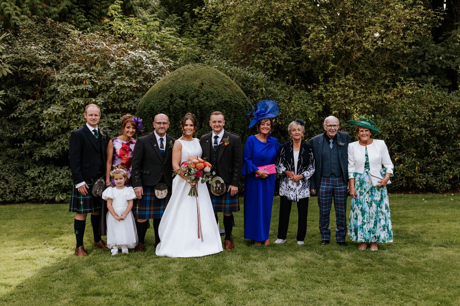 Large wedding group portrait with mixed formal attire and kilts against garden landscaping.