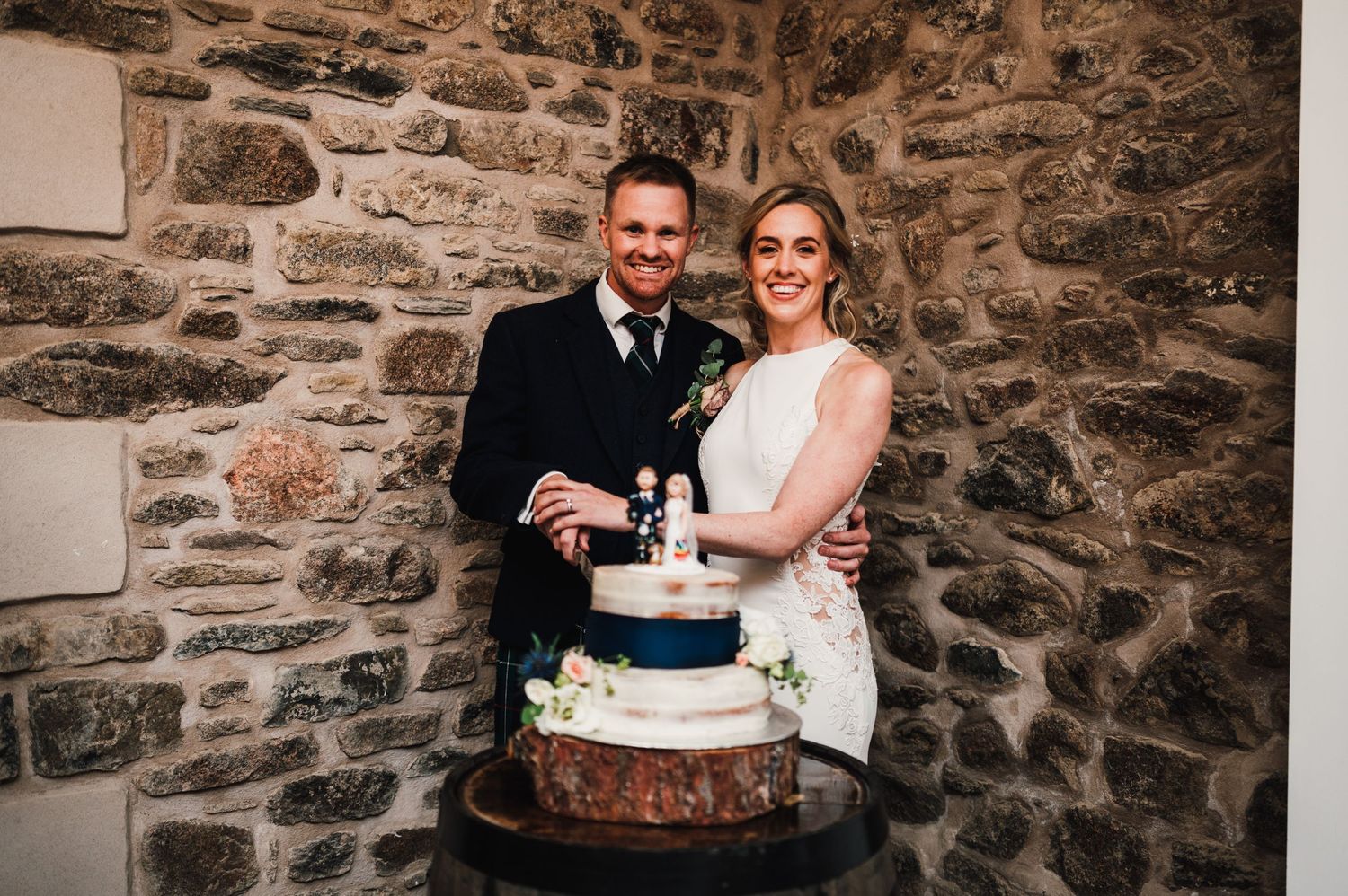 Wedding couple posing with their rustic three-tier cake against a stone wall backdrop.