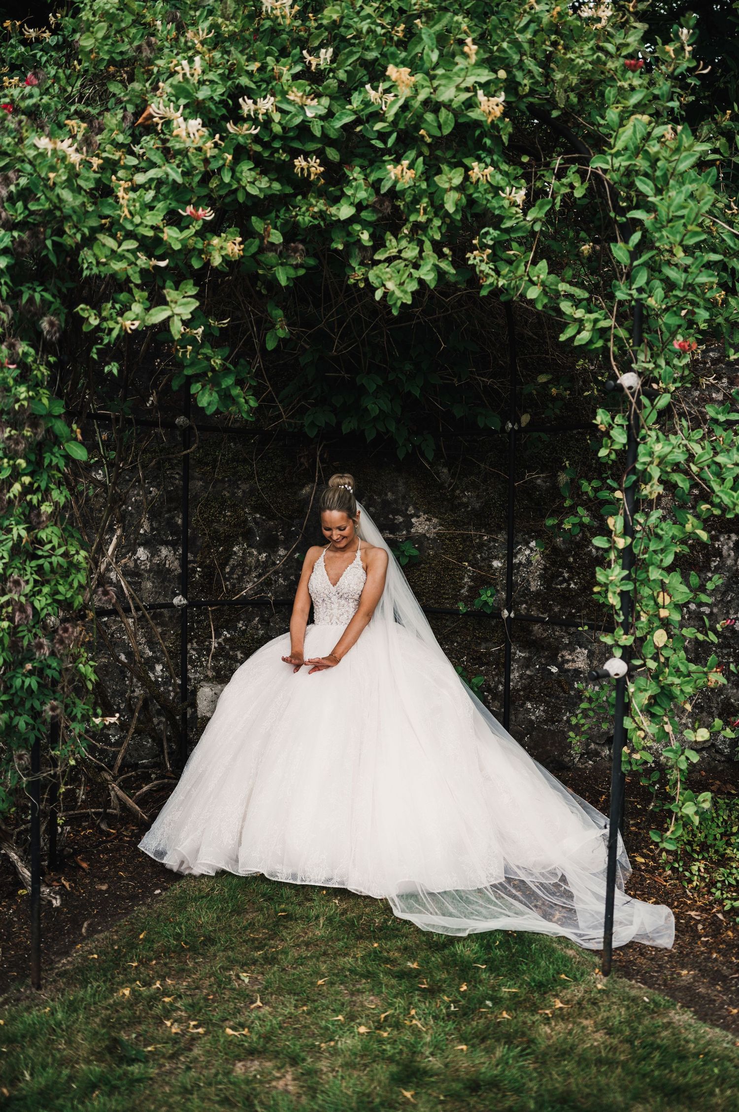 Bride in elegant white ballgown poses against stone wall surrounded by climbing vines and foliage.