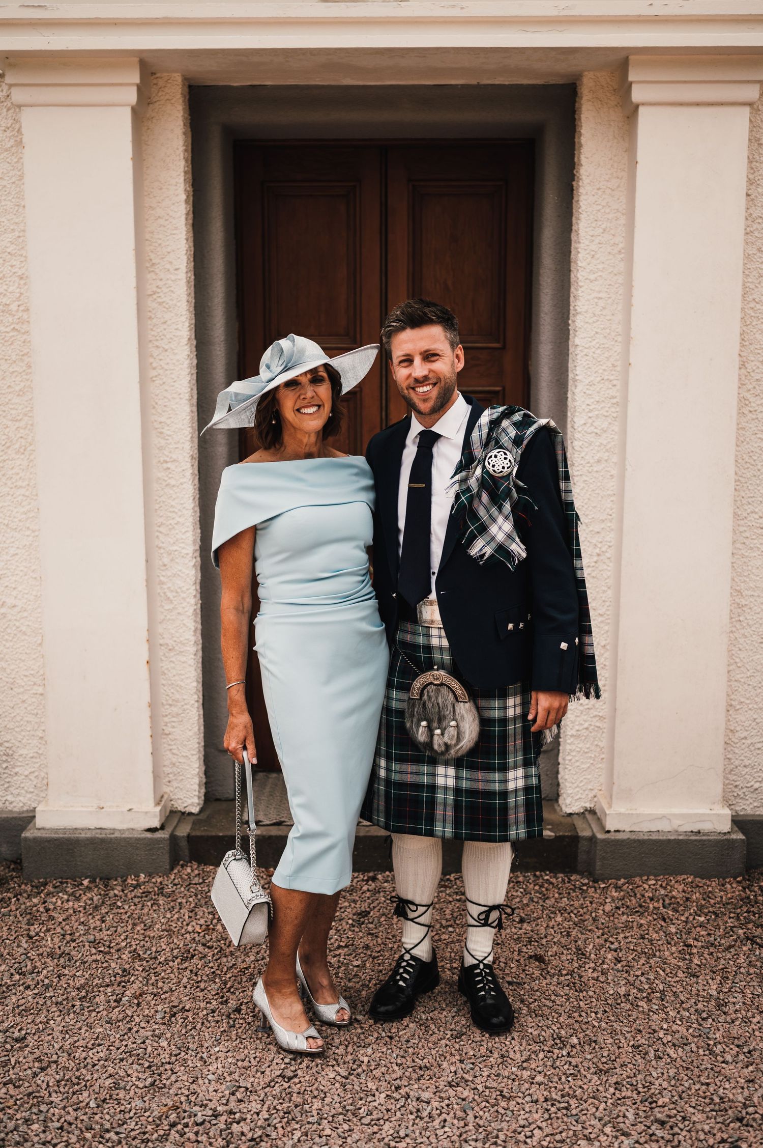 Couple in formal attire poses by doorway, with man wearing traditional Scottish kilt and woman in blue dress.