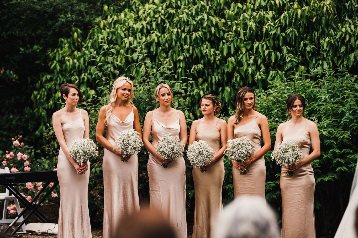 Row of bridesmaids in blush pink dresses holding bouquets at outdoor ceremony.